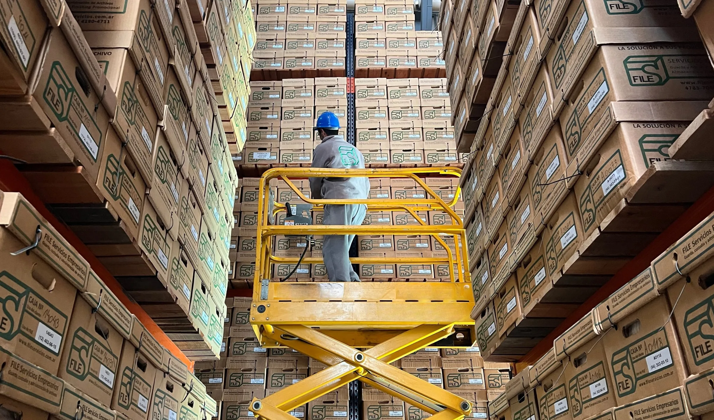 Operario sobre una plataforma elevadora entre estanterías de cajas del depósito