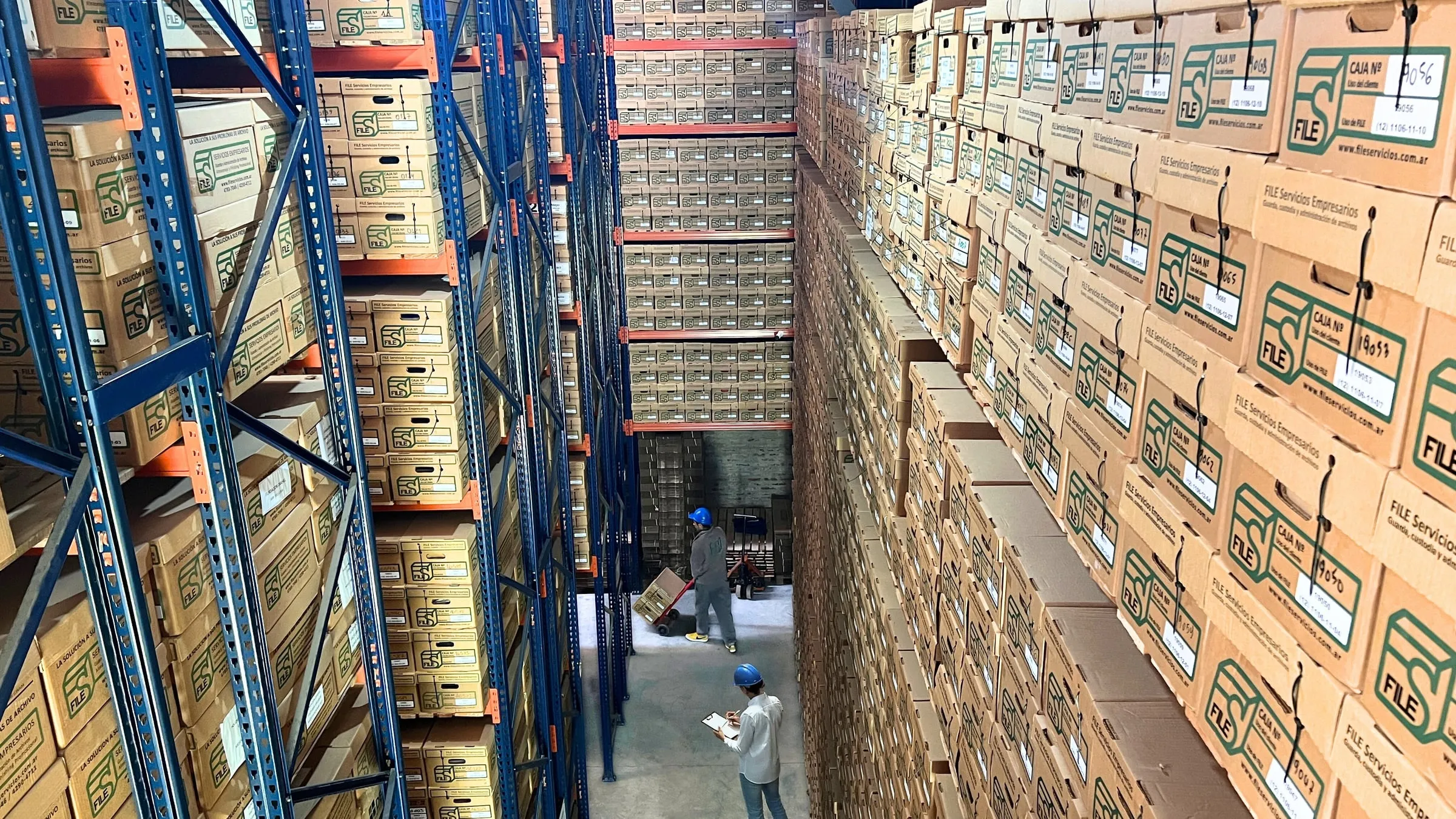 Vista general del depósito de FILE: pasillo entre estanterías de cajas numeradas, con dos operarios trabajando